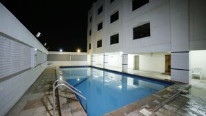 Una piscina con bordo piastrellato, scala in metallo e un edificio bianco sullo sfondo.