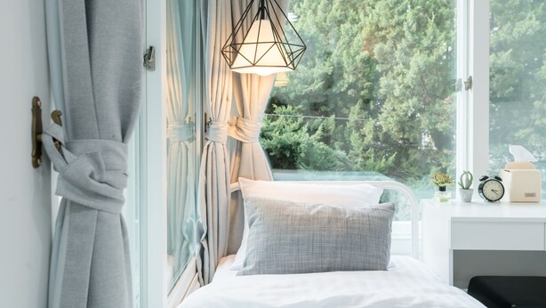 Un dormitorio con cama, ventana con cortinas, mesita de noche con reloj y vista a árboles desde el exterior.