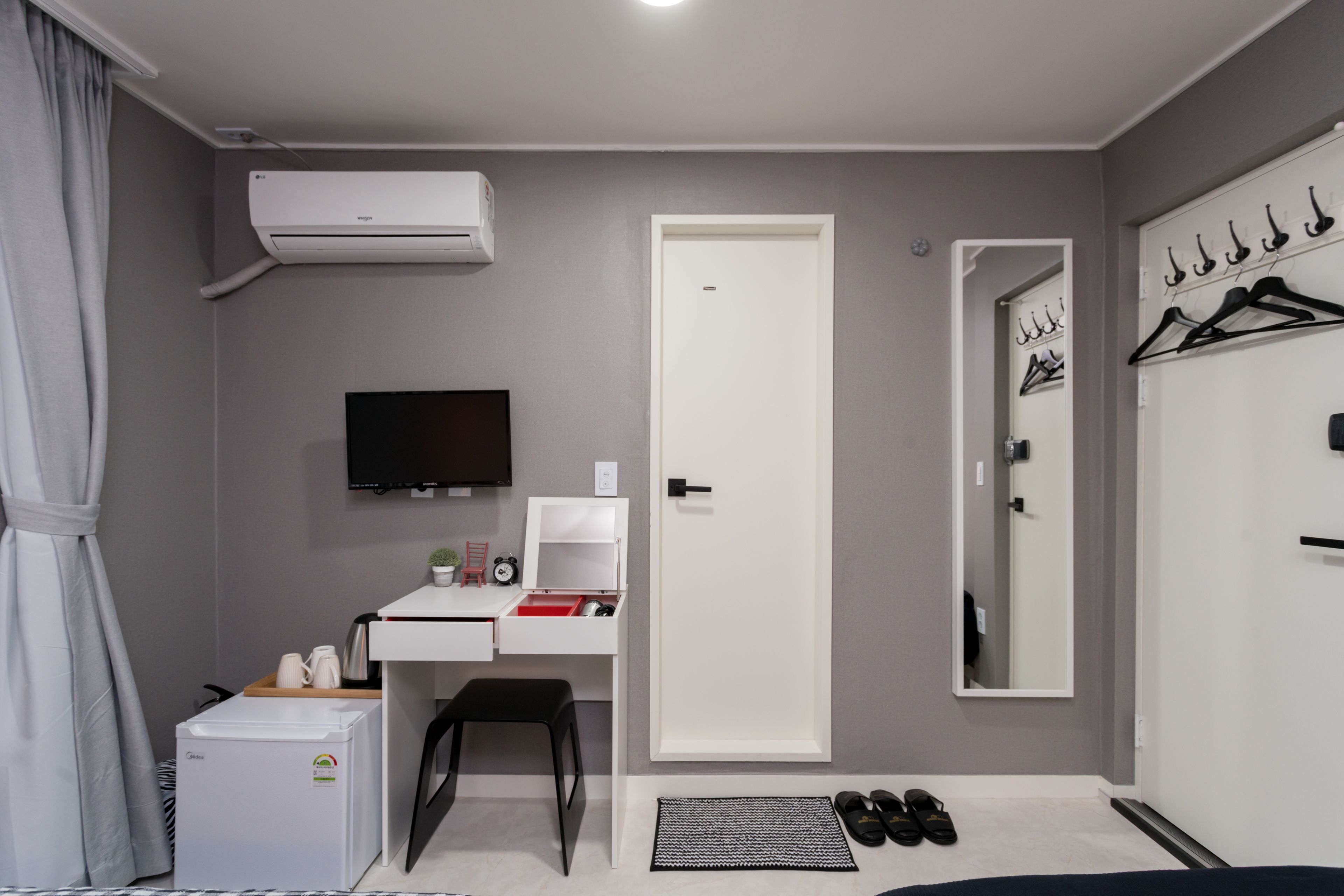 A compact bedroom with a bed, a desk with a chair, a TV, a wardrobe with hangers, a mirror, and a radiator.