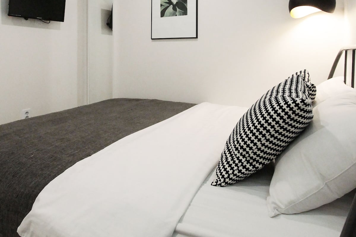 Un dormitorio con cama, un cojín blanco y negro estampado, un cuadro enmarcado, un televisor montado y una planta colgante.