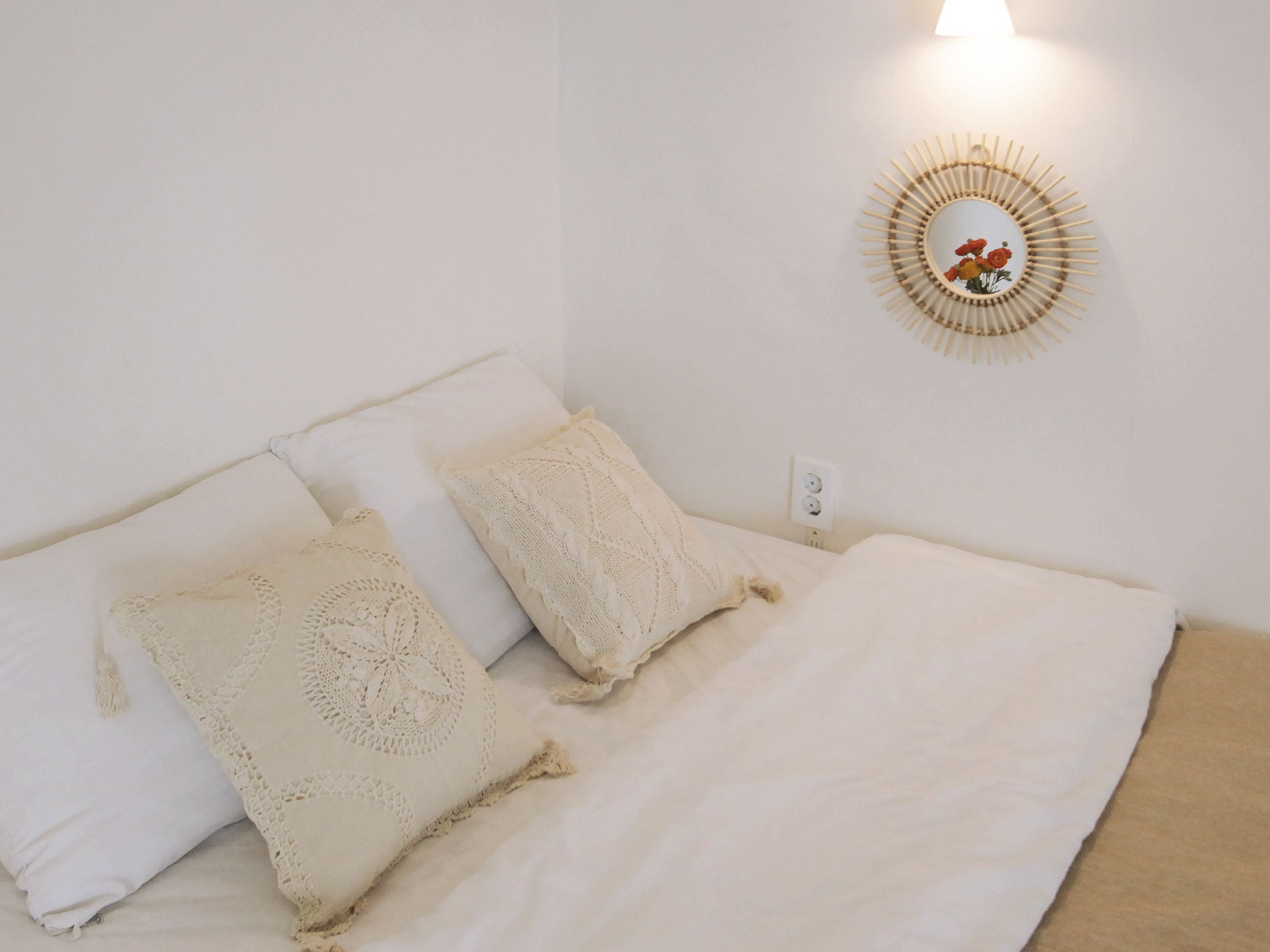 Een netjes opgemaakt bed met witte lakens en twee decoratieve kussens, een wandlamp en een ronde spiegel met een bloemistisch ontwerp.