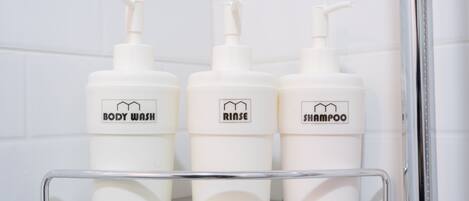 Tres botellas blancas de jabón corporal, aclarador y champú sobre una repisa metálica en un baño con azulejos blancos.