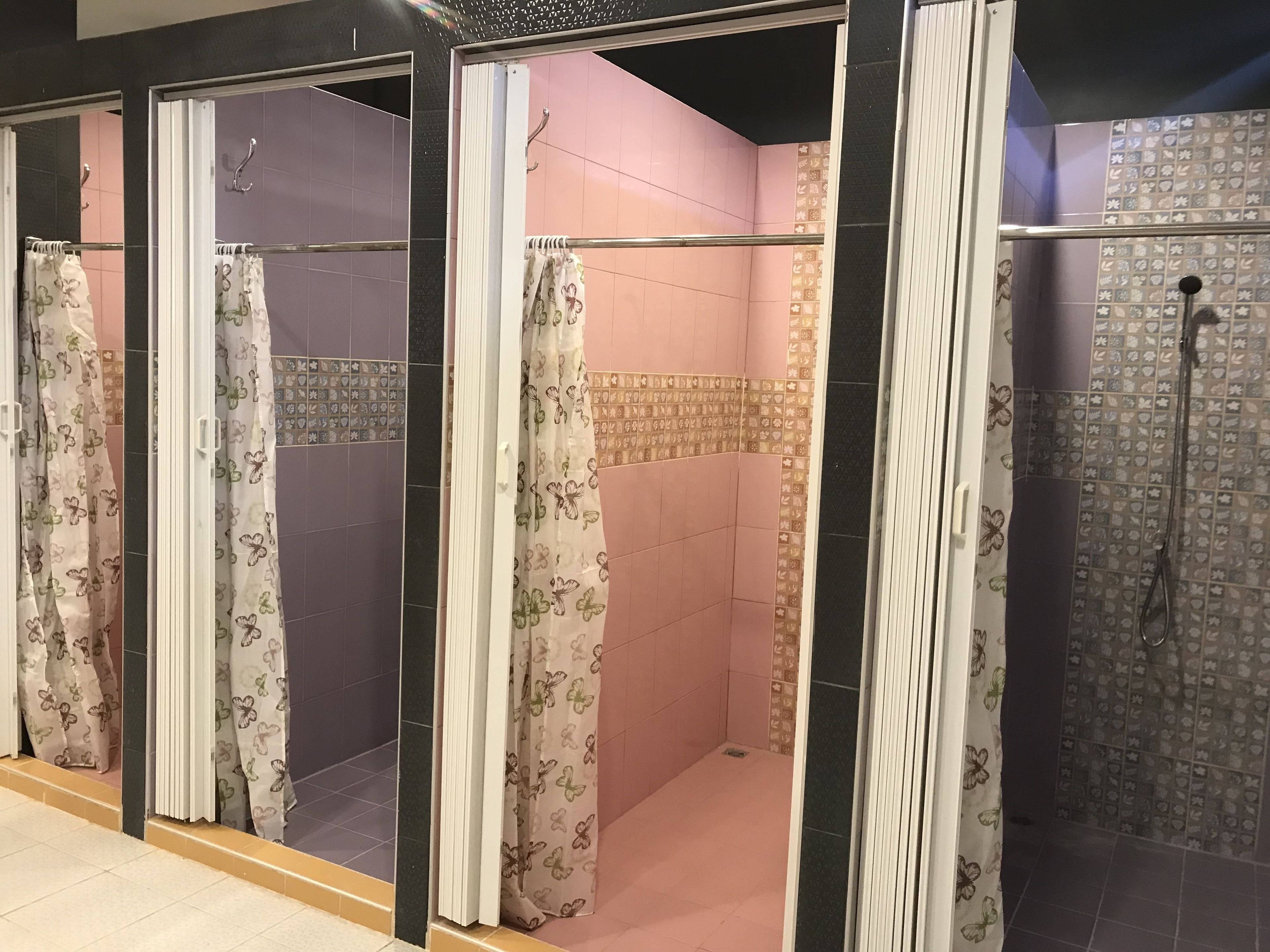 Una hilera de cabinas de ducha con cortinas estampadas con motivos de mariposas, paredes de azulejos en tonos rosa y morado, y una zona de ducha con pared de azulejos y cabezal de ducha.