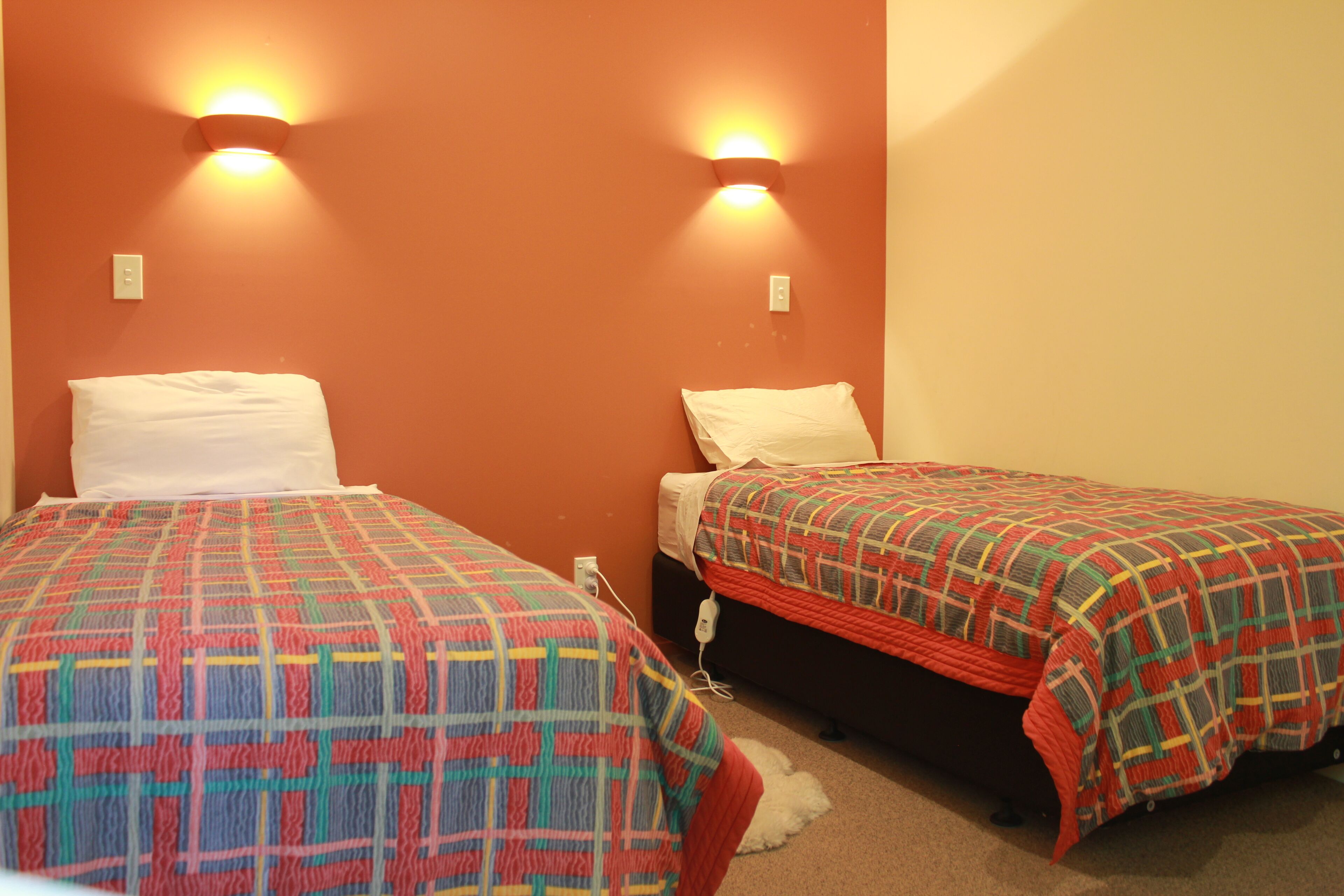 Two single beds with patterned bedding, a wall-mounted light fixture, and a power outlet on the wall.