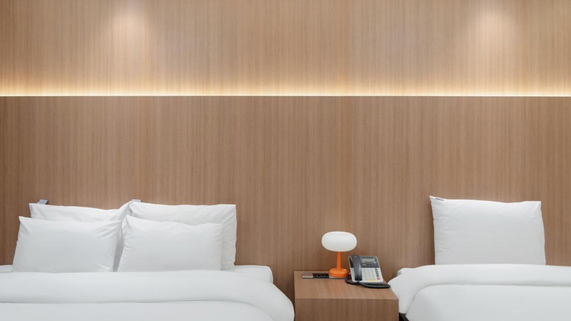 A hotel room with a wooden headboard, two beds with white bedding, a bedside table with a lamp and phone, and a wall-mounted light fixture.