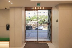 Entrada de hotel con puerta de cristal, felpudo verde y zona de estacionamiento visible afuera.