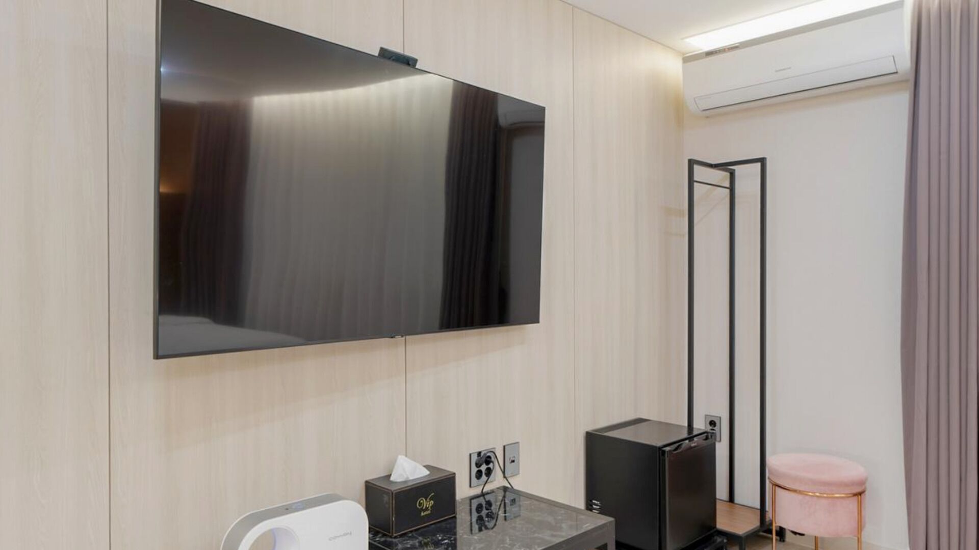 A modern hotel room with a large flat-screen TV, a compact black refrigerator, a marble countertop, and a pink cushioned stool.