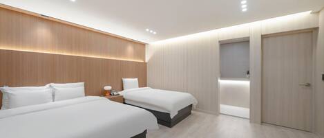 A modern hotel room with two beds, a wooden headboard, a bedside table with a lamp, and a door leading to a bathroom.