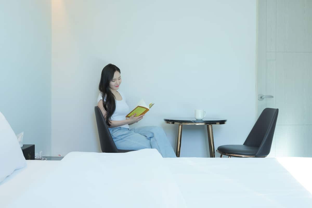 Una mujer sentada en una cama de un cuarto de hotel, leyendo un libro. Hay una mesita redonda con una taza blanca y una silla negra junto a la cama.