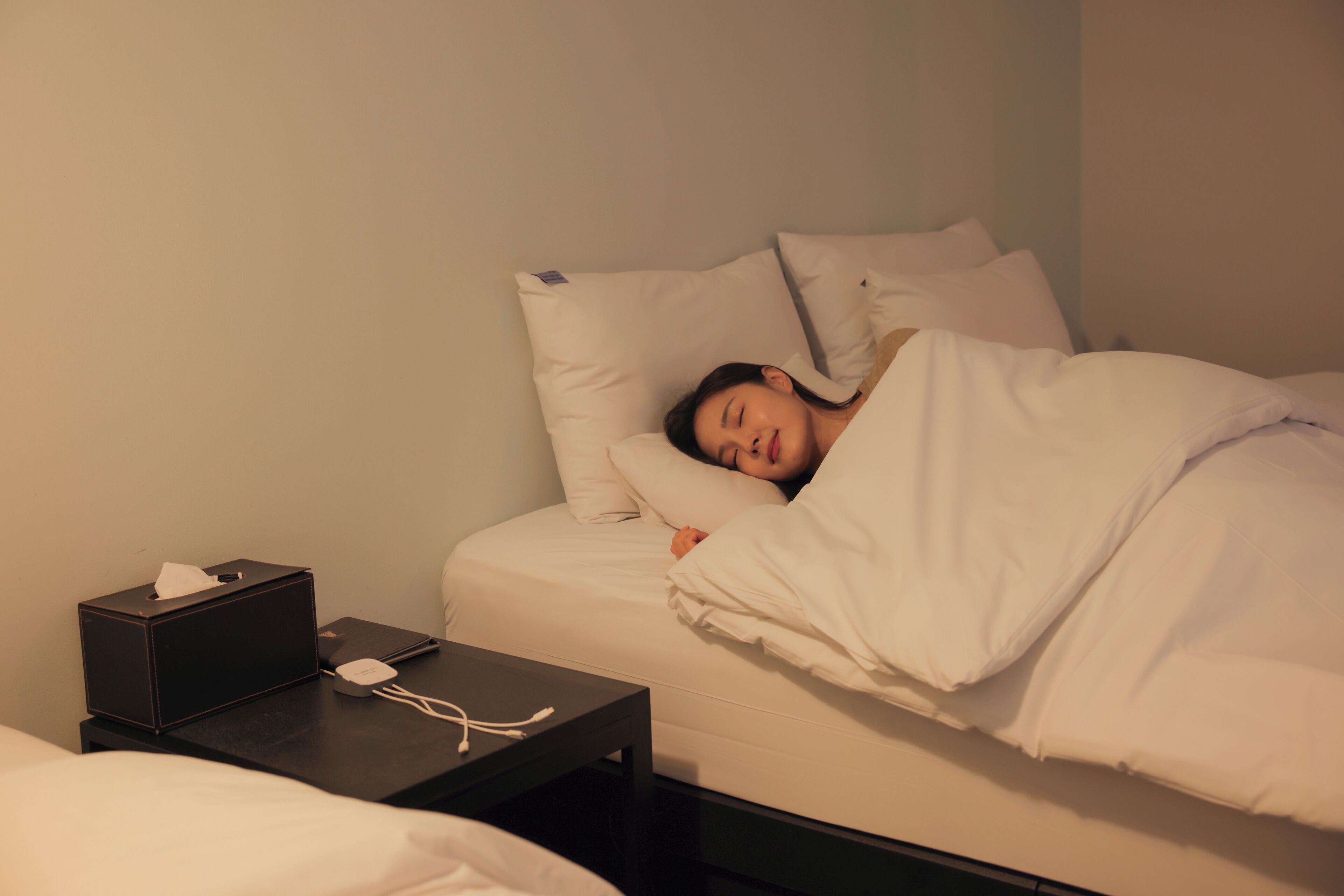 A person sleeping in a bed with white bedding, a black bedside table with a tissue box and a phone charger, and a plain wall in the background.