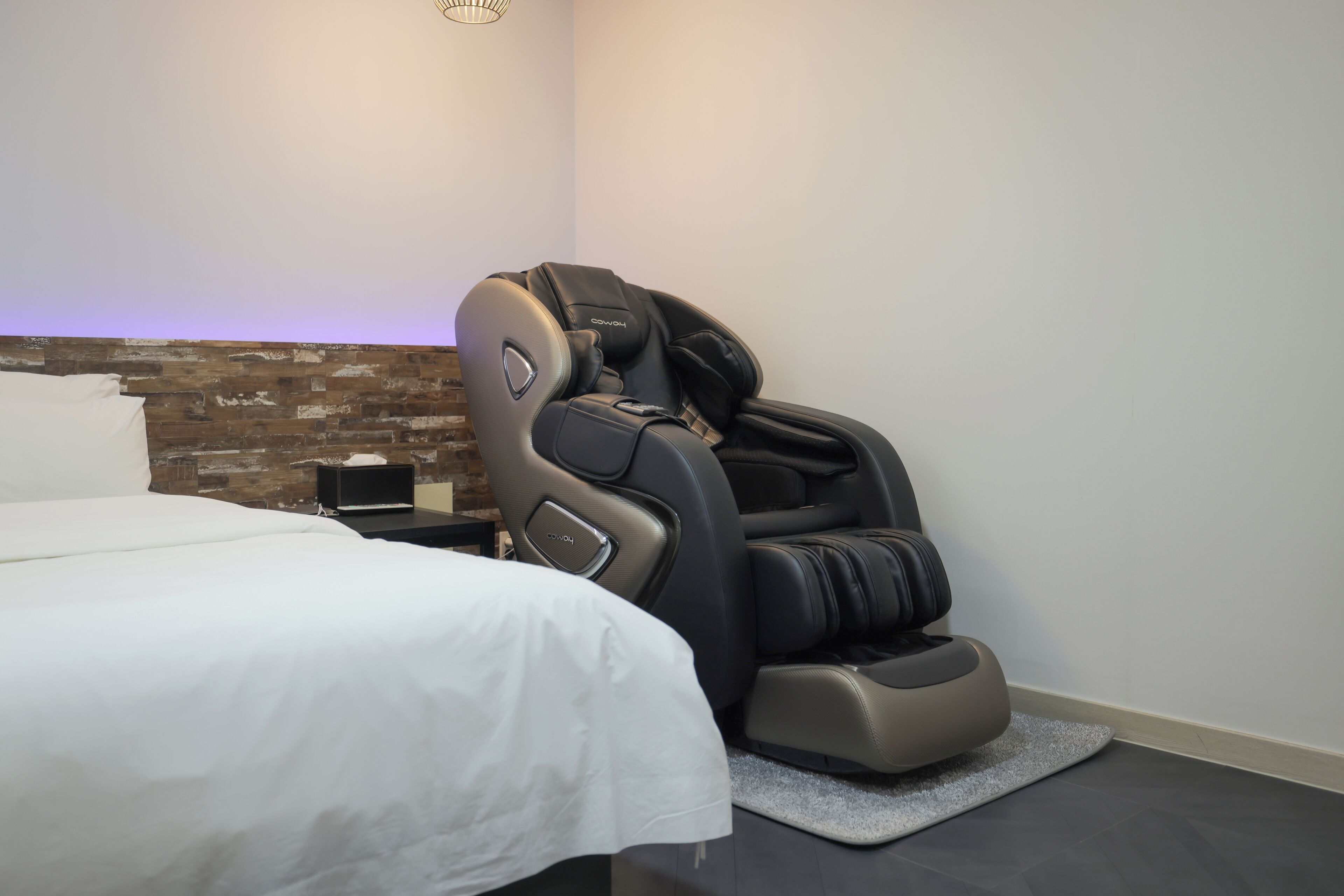 A massage chair in a room with a bed, a small table, and a brick-patterned wall.
