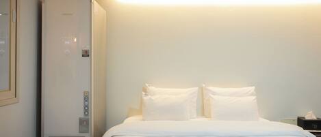 Habitación de hotel con cama, ropa de cama blanca, una mesita de noche, una lámpara y un calentador de pared.