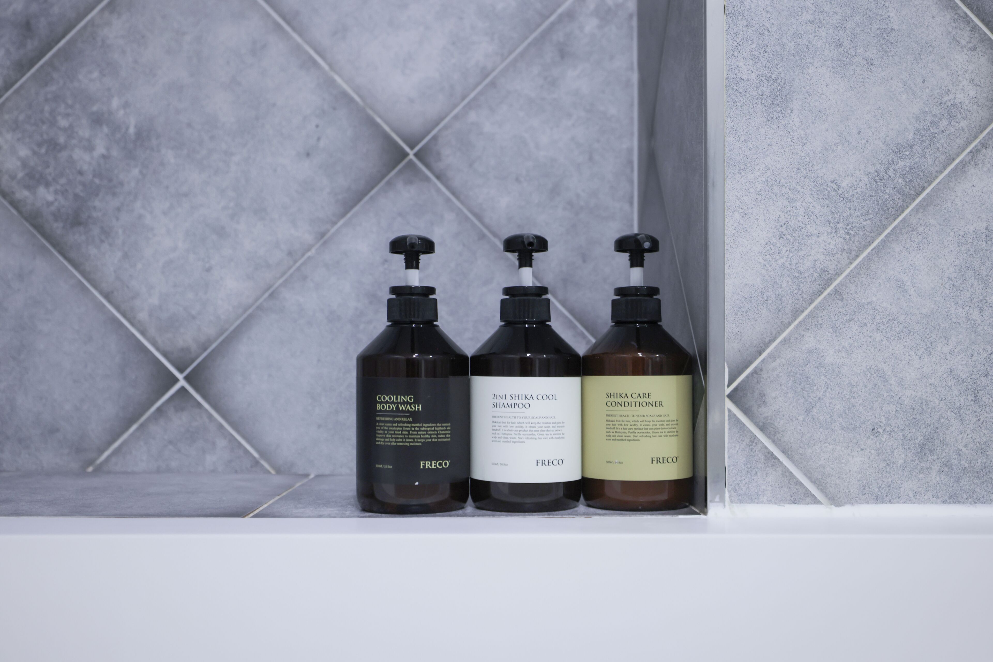 Three bottles of Freco body wash and conditioner on a shelf against a tiled wall.