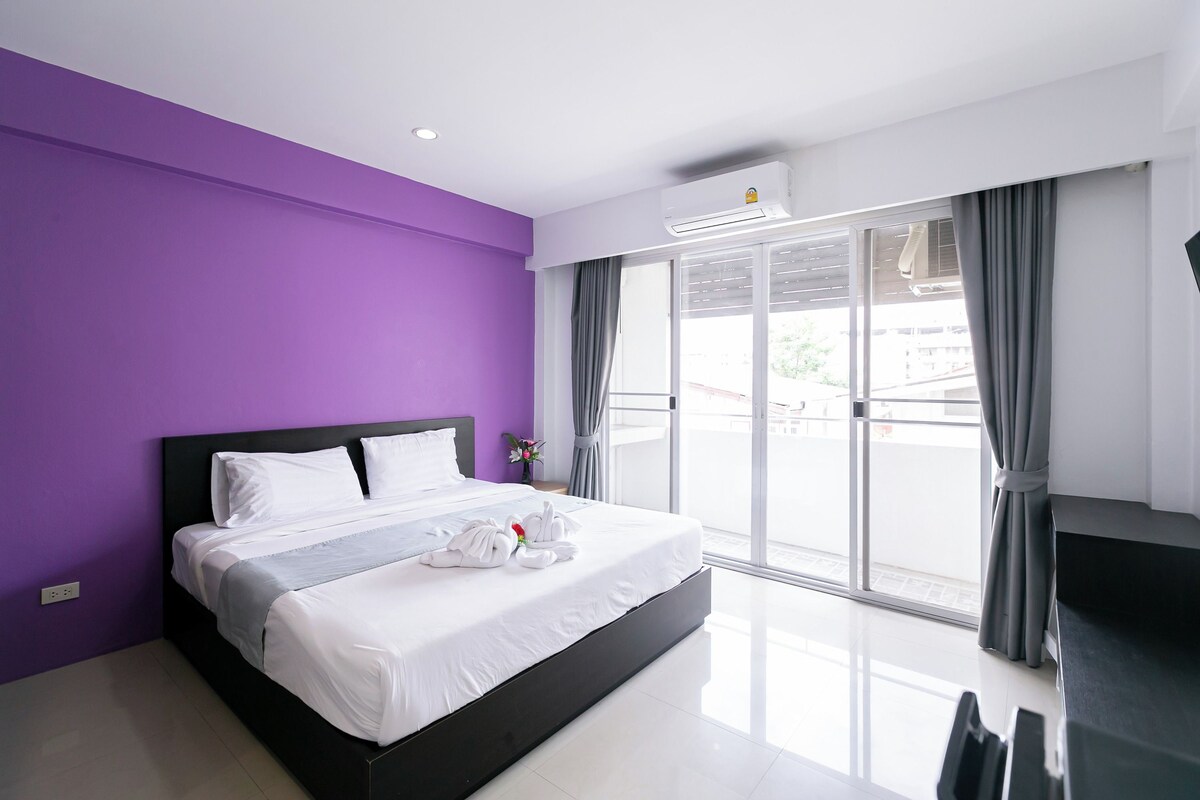 A modern hotel room with a large bed, purple walls, and a view of the outside.