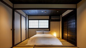 A traditional Japanese-style bedroom with tatami flooring, a bed with white bedding, a window with vertical bars, and a sliding door.