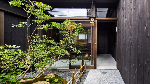 A traditional Japanese-style entrance with a wooden gate, a small garden with rocks and bamboo, and a clear pathway leading inside.