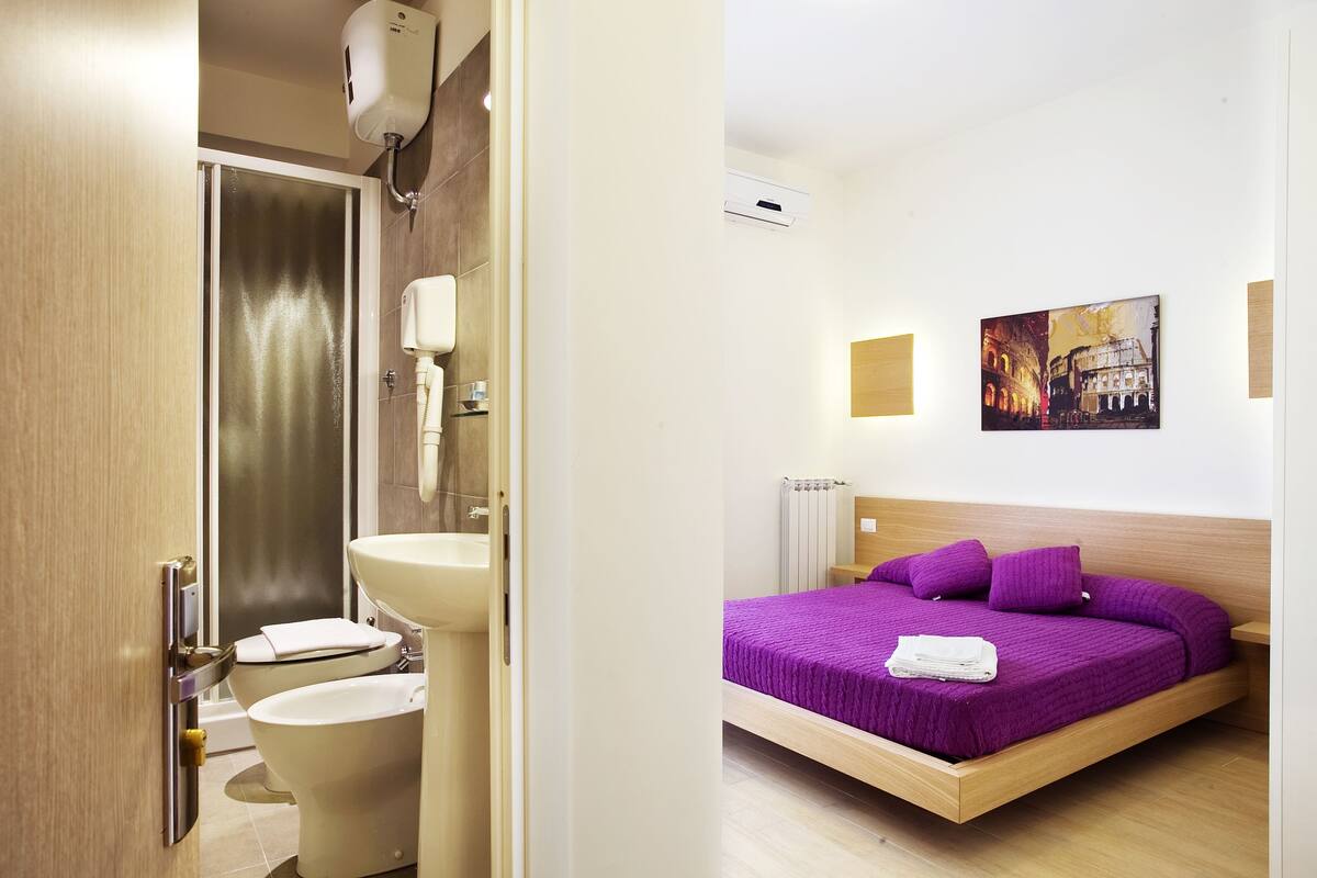 Un baño moderno con ducha, inodoro y lavabo. La habitación tiene suelo de madera, una cama con colcha púrpura y un aire acondicionado de pared.