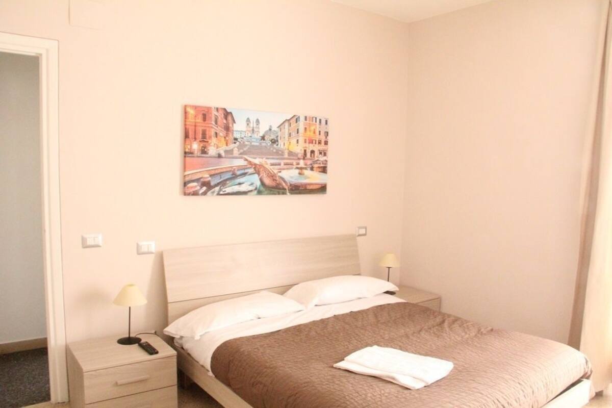 Una habitación de hotel con una cama, una mesita de noche con una lámpara, un cuadro de un paisaje urbano y una puerta.