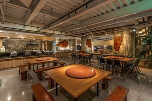 Un restaurante espacioso con techos altos, vigas a la vista y una combinación de piedra natural y elementos de madera. Mesas y sillas están dispuestas para comer, y se observa una barra con un menú.