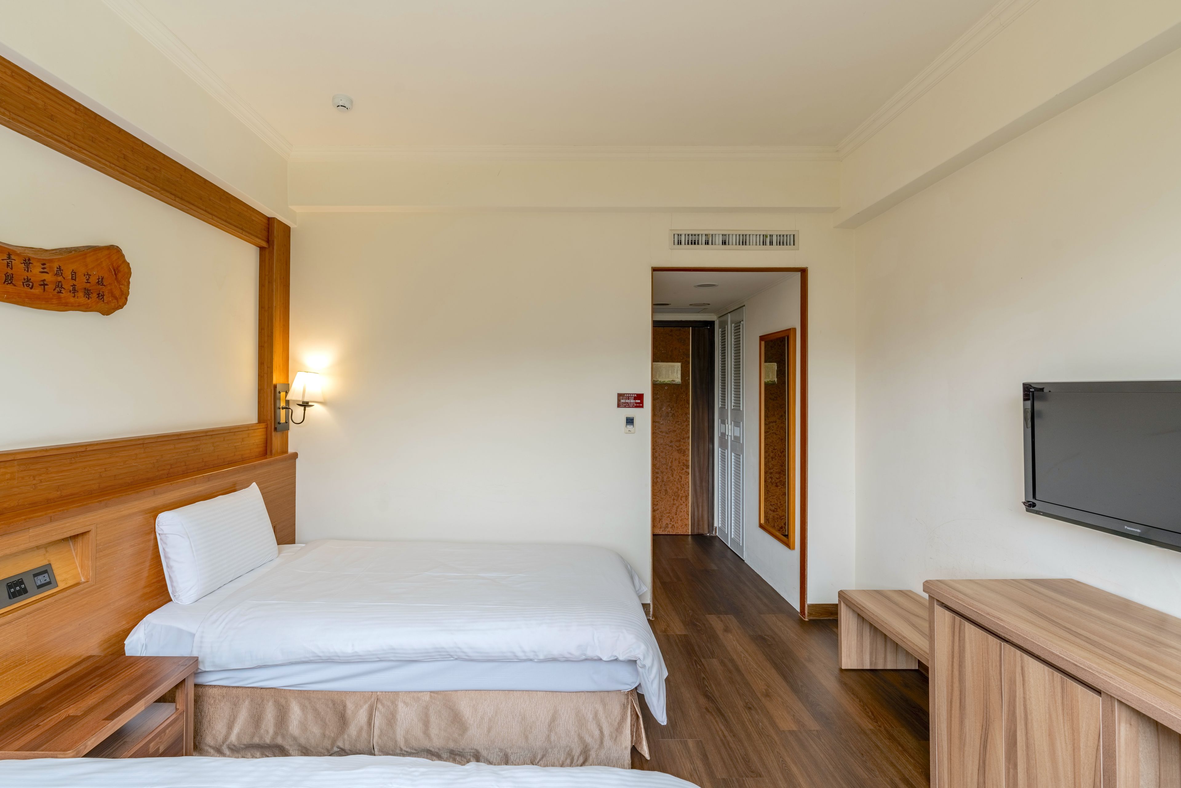 A hotel room with two beds, a wooden headboard, a wall-mounted TV, and a wooden bench.
