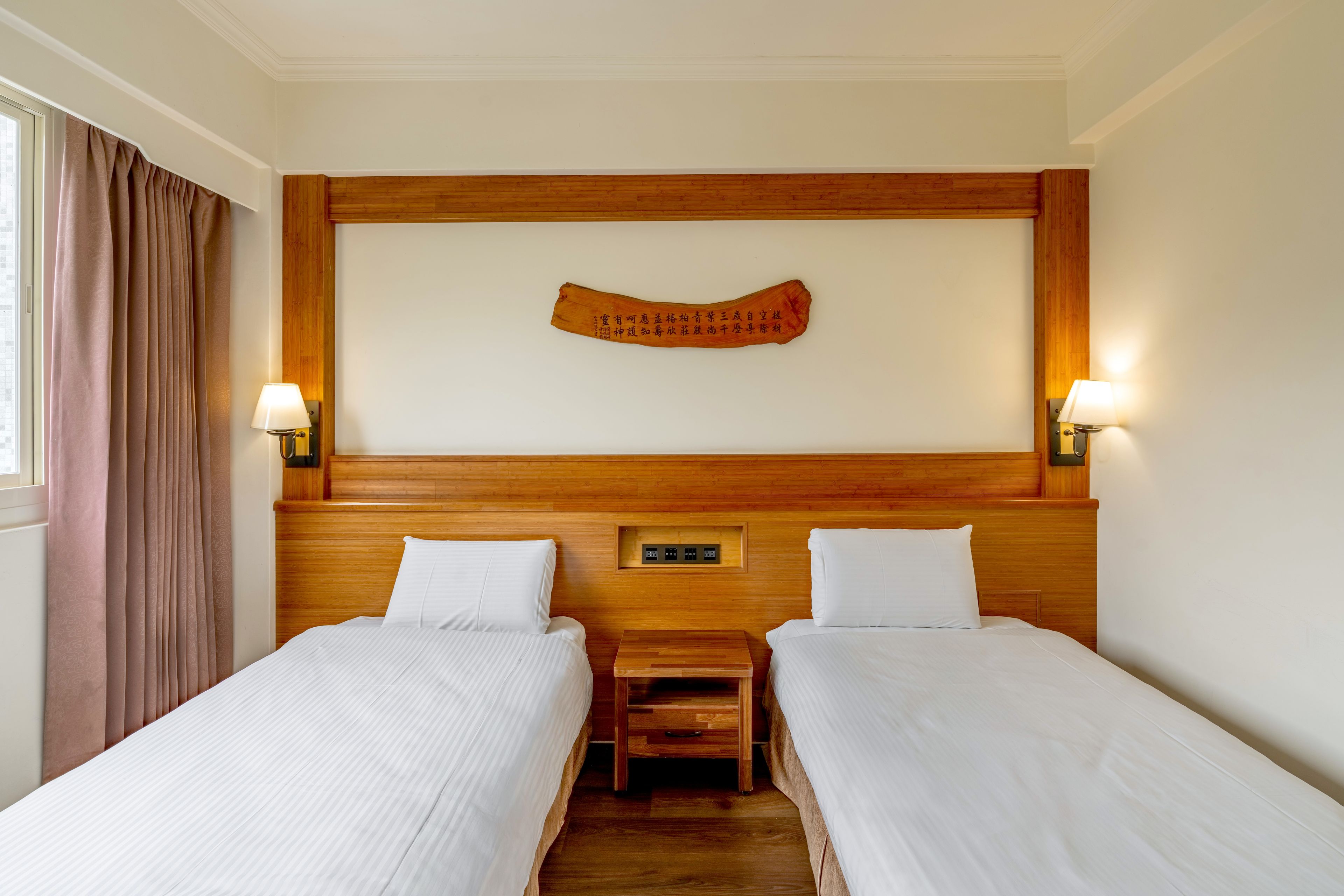 A hotel room with two beds, a wooden headboard, a wall-mounted lamp, and a nightstand.