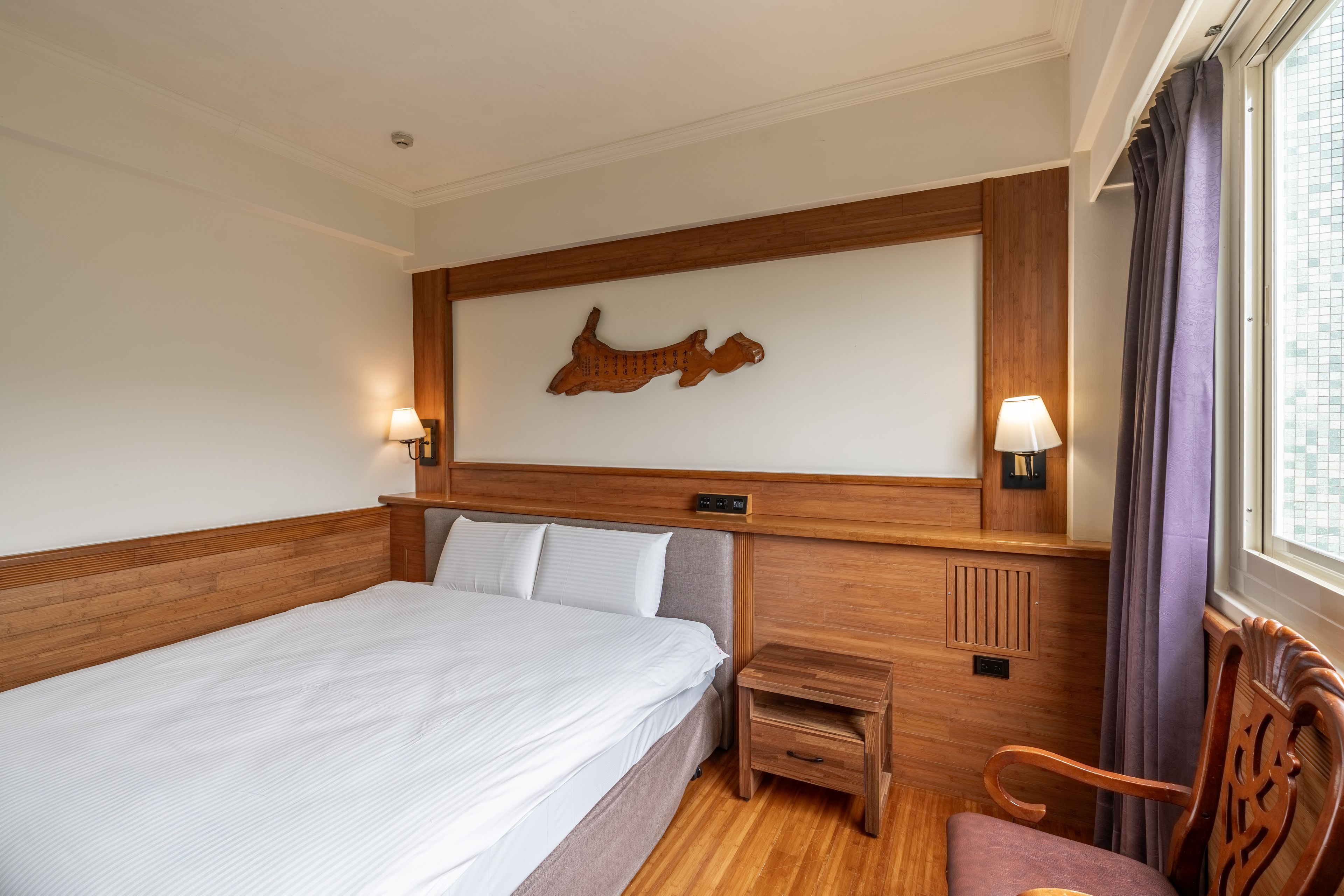 A hotel room with a large bed, wooden headboard, two bedside lamps, a nightstand, a chair, and a wall-mounted artwork.
