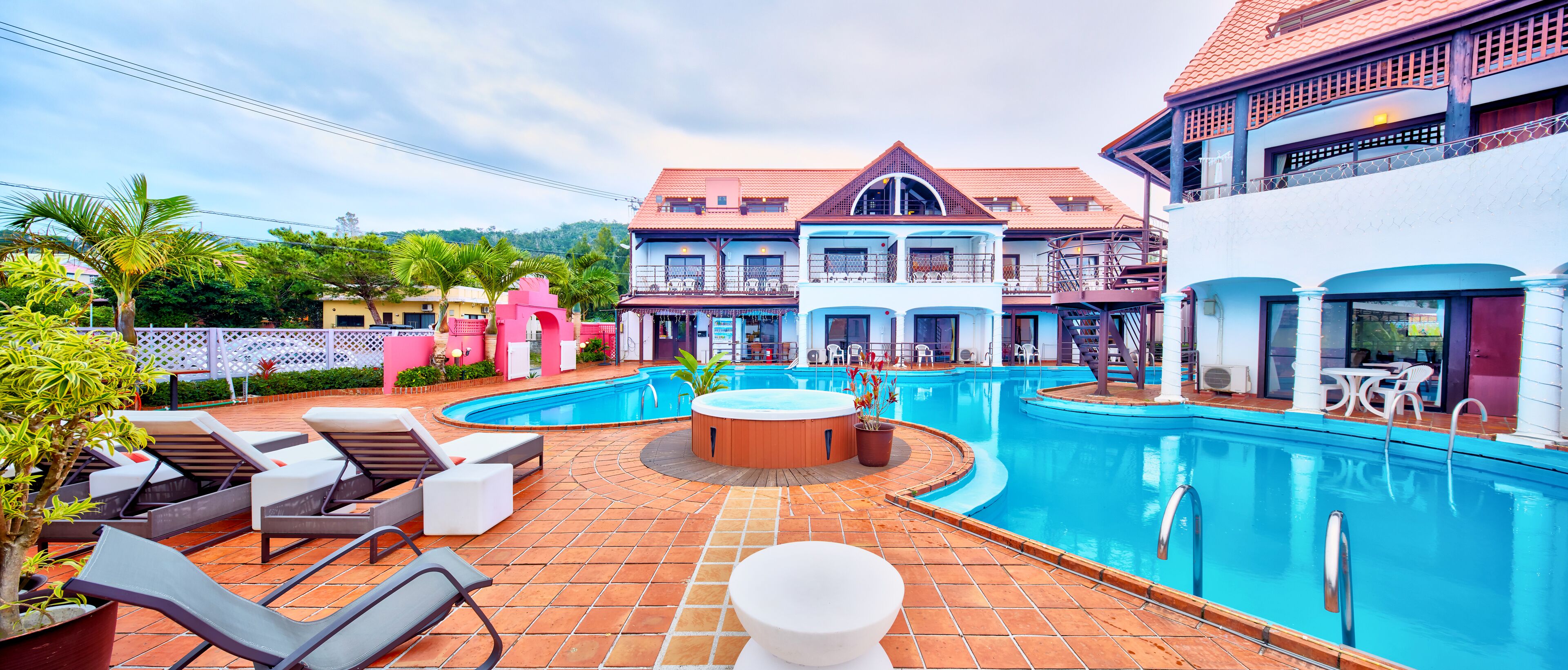 A swimming pool area with lounge chairs, a hot tub, and a view of a large building with balconies.