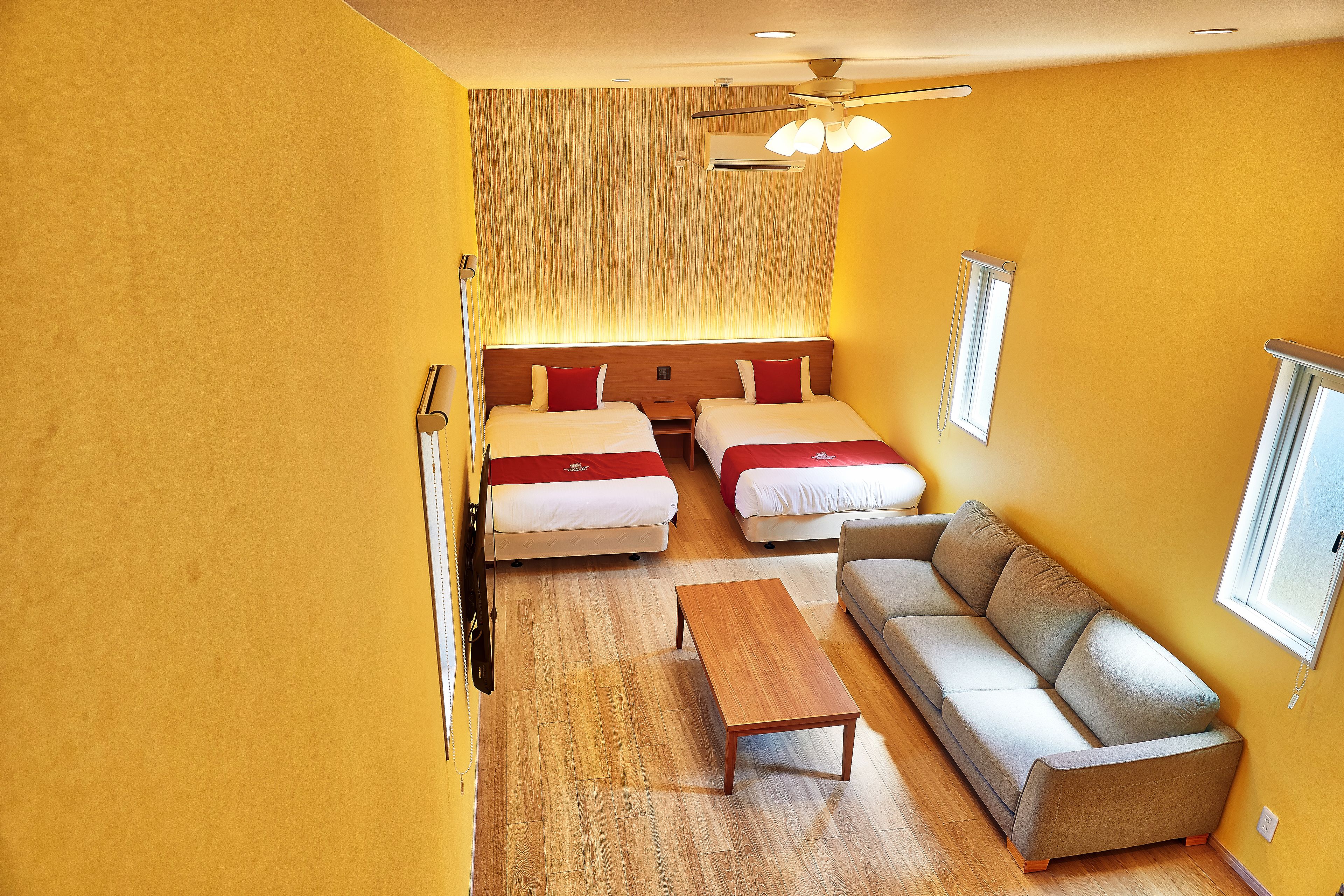 A hotel room with two single beds, a sofa, a wooden coffee table, and a ceiling fan.