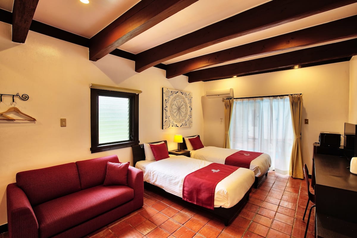 A hotel room with two beds, a red sofa, a wooden ceiling, and a window with curtains.