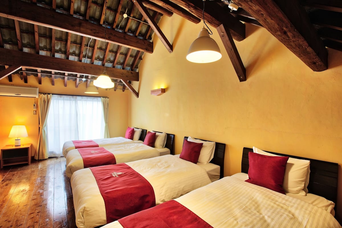 A hotel room with three beds, wooden beams, a ceiling fan, and a window with curtains.