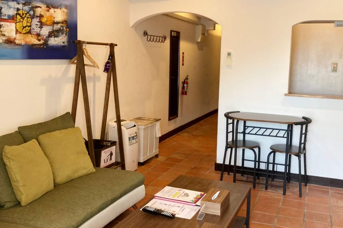 A living room with a green sofa, a small table with chairs, a painting on the wall, and a door leading to another room.