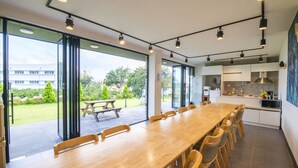 Una cocina moderna con una isla grande, un comedor con una mesa larga y sillas, y vista a un jardín a través de ventanas amplias.