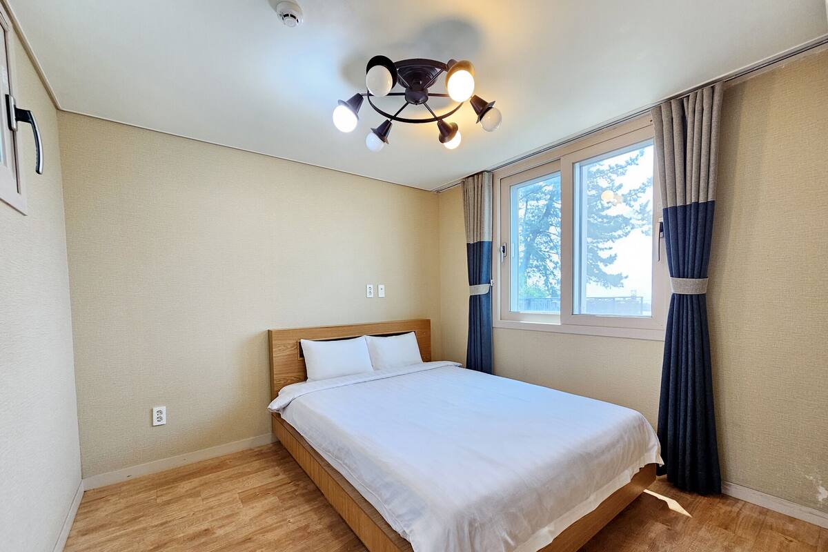 A bedroom with a wooden bed, a ceiling light fixture, a window with curtains, and a view of trees outside.