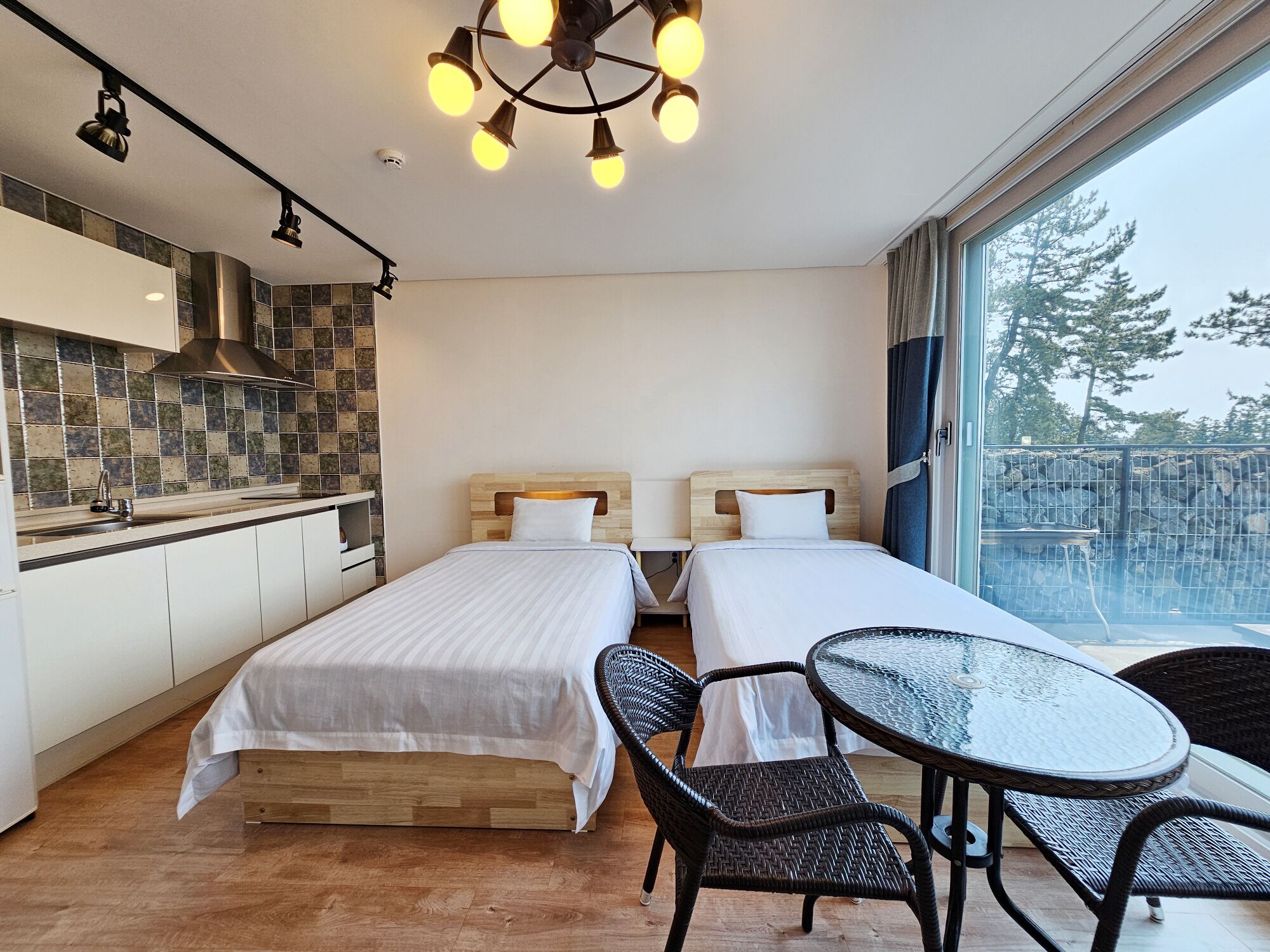 A modern hotel room with two beds, a kitchen area, and a balcony with a view of trees.
