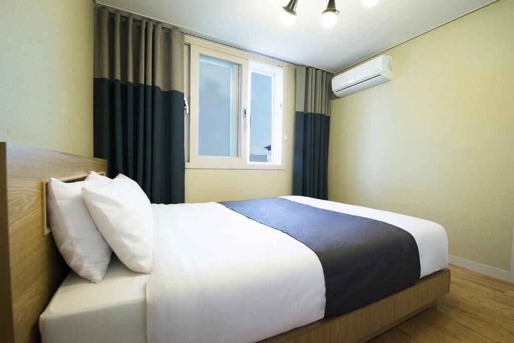 A neatly made bed with white and dark bedding, a window with curtains, and an air conditioning unit on the wall.