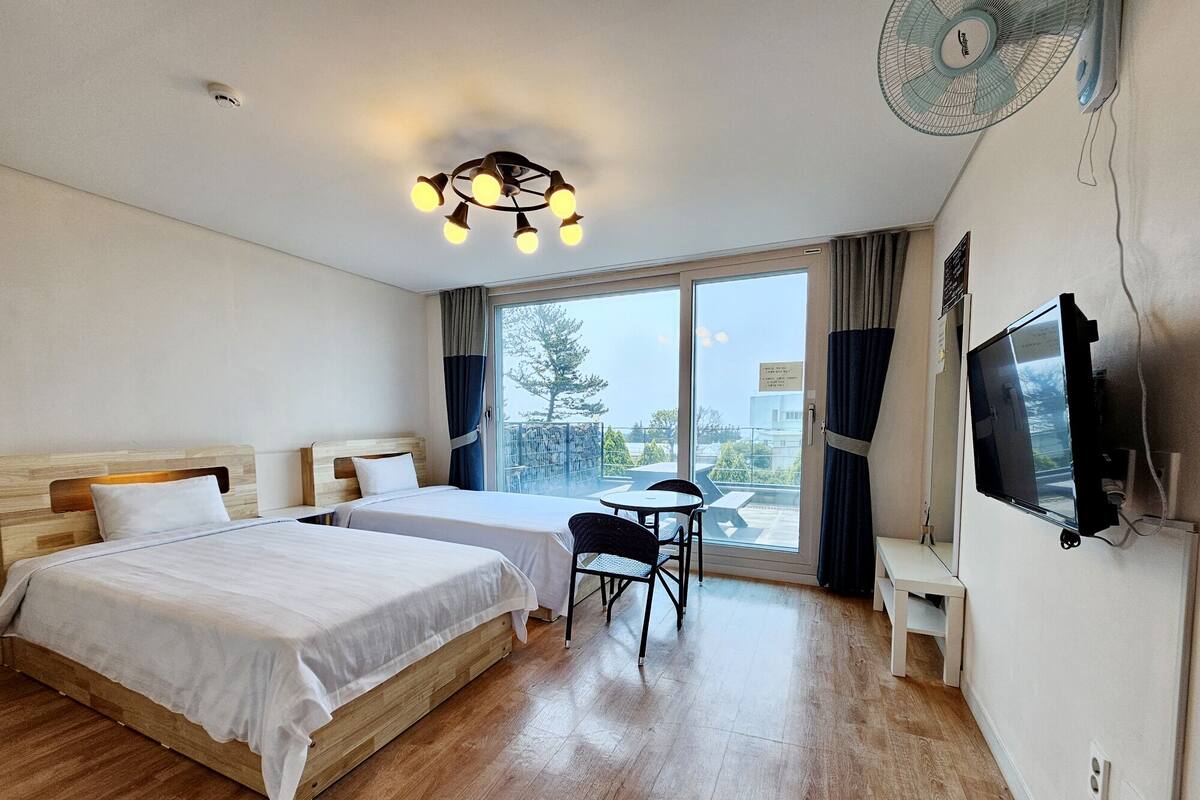 A hotel room with two beds, a desk, a chair, a TV, and a large window with curtains.