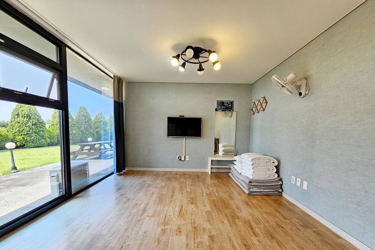 A spacious room with a large window, wooden flooring, a flat-screen TV, a mirror, and a stack of towels.