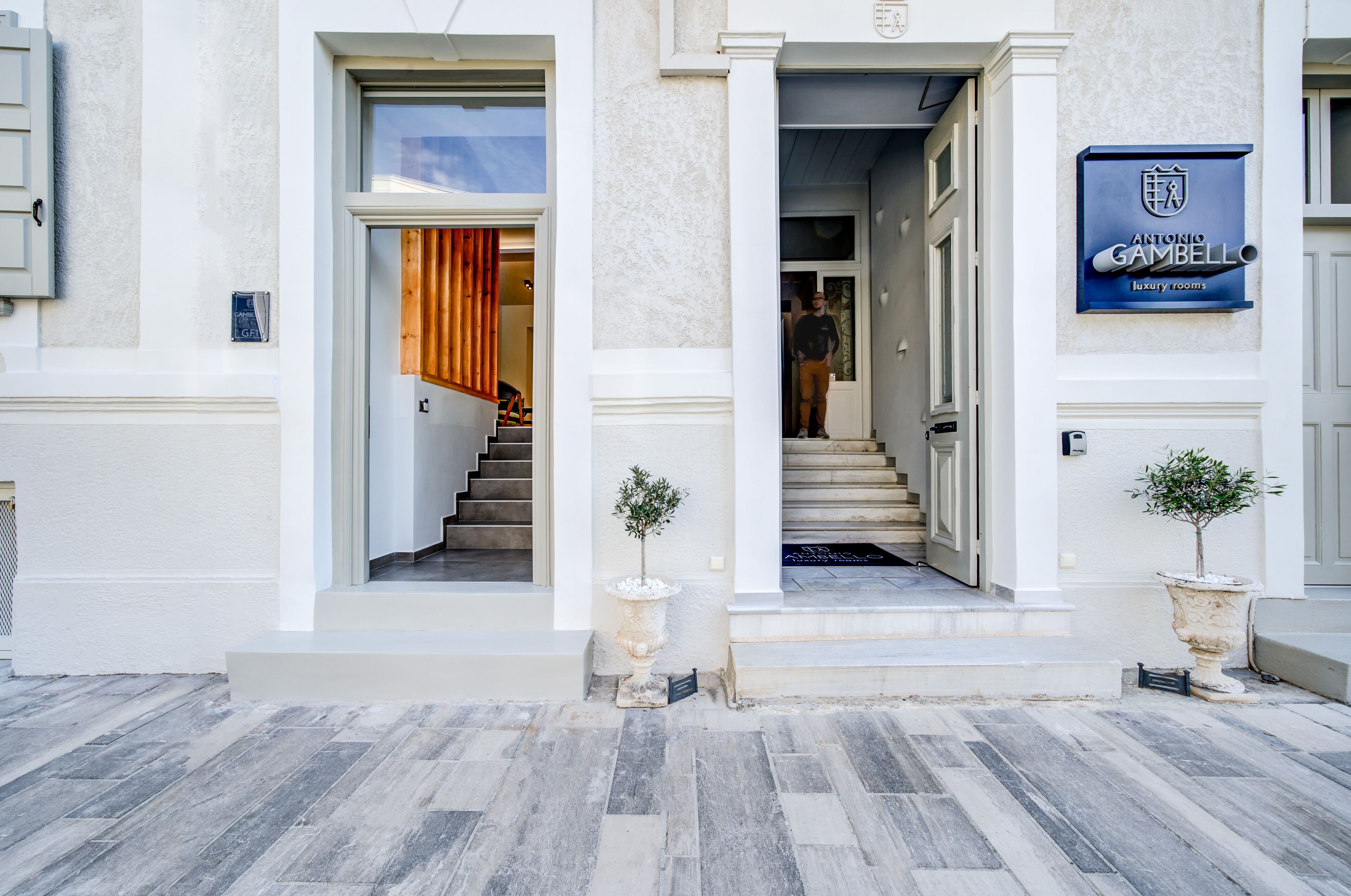 A building entrance with a sign for Antonio Gambello luxury rooms, a staircase, and two potted plants.