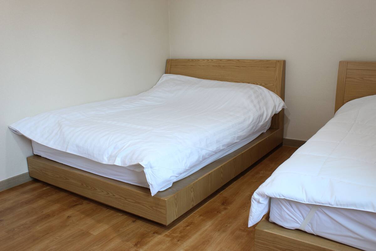 Una cama individual con estructura de madera y ropa de cama blanca en una habitación con suelo de madera y paredes lisas.