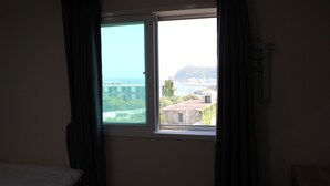 A room with a single bed, a window with curtains, and a view of buildings and a body of water. - The Lucete Hotel (Seogwipo)