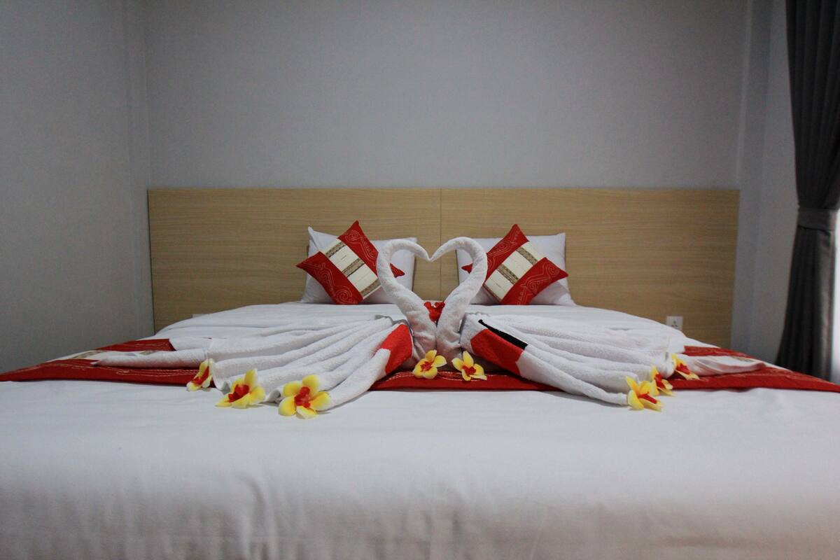 Cama bem arrumada com toalhas dispostas em formato de coração e algumas flores sobre ela.