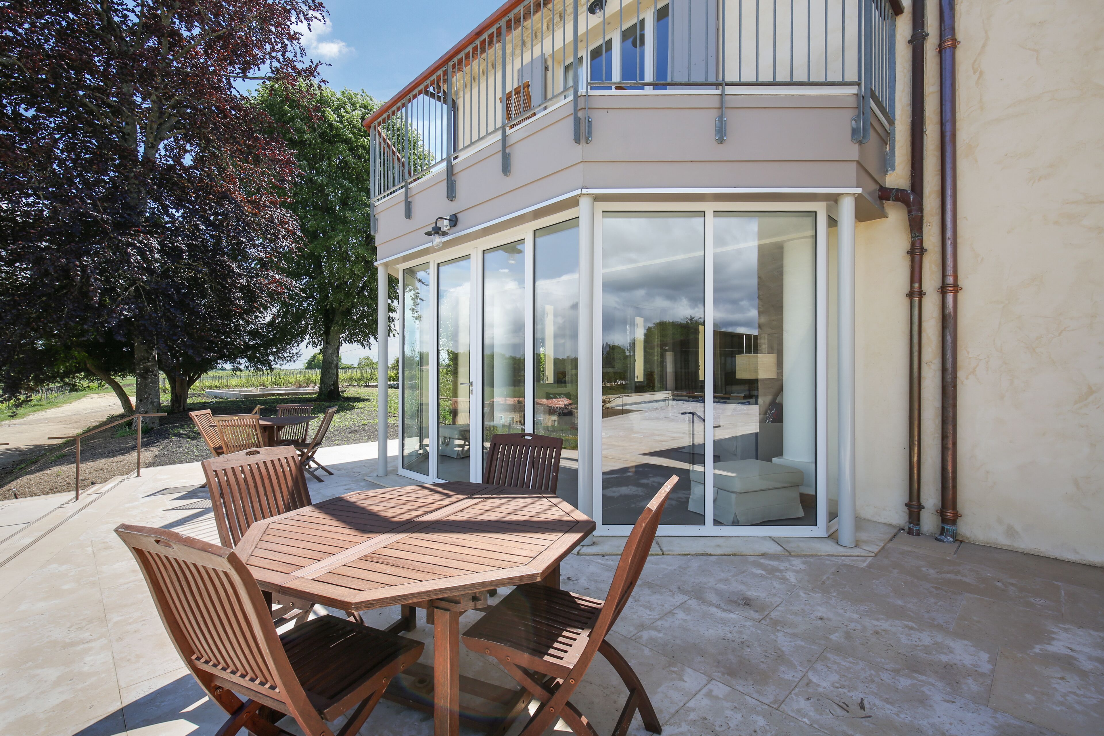 Un patio al aire libre con mesa y sillas de madera, un ventanal de vidrio que da a un balcón y un árbol al fondo.