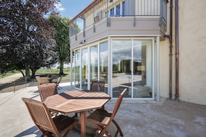 Un patio al aire libre con mesa y sillas de madera, un ventanal de vidrio que da a un balcón y un árbol al fondo.