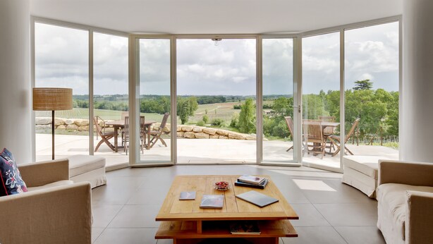 A spacious room with a large glass door leading to an outdoor area, a wooden table with books, and a view of a green landscape. - Logis DE Valandraud (Saint-Etienne-De-Lisse)