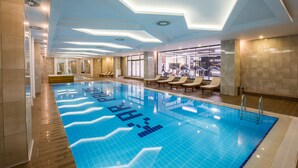 Una piscina interior moderna con luces de techo geométricas, paredes de vidrio transparente y tumbonas.