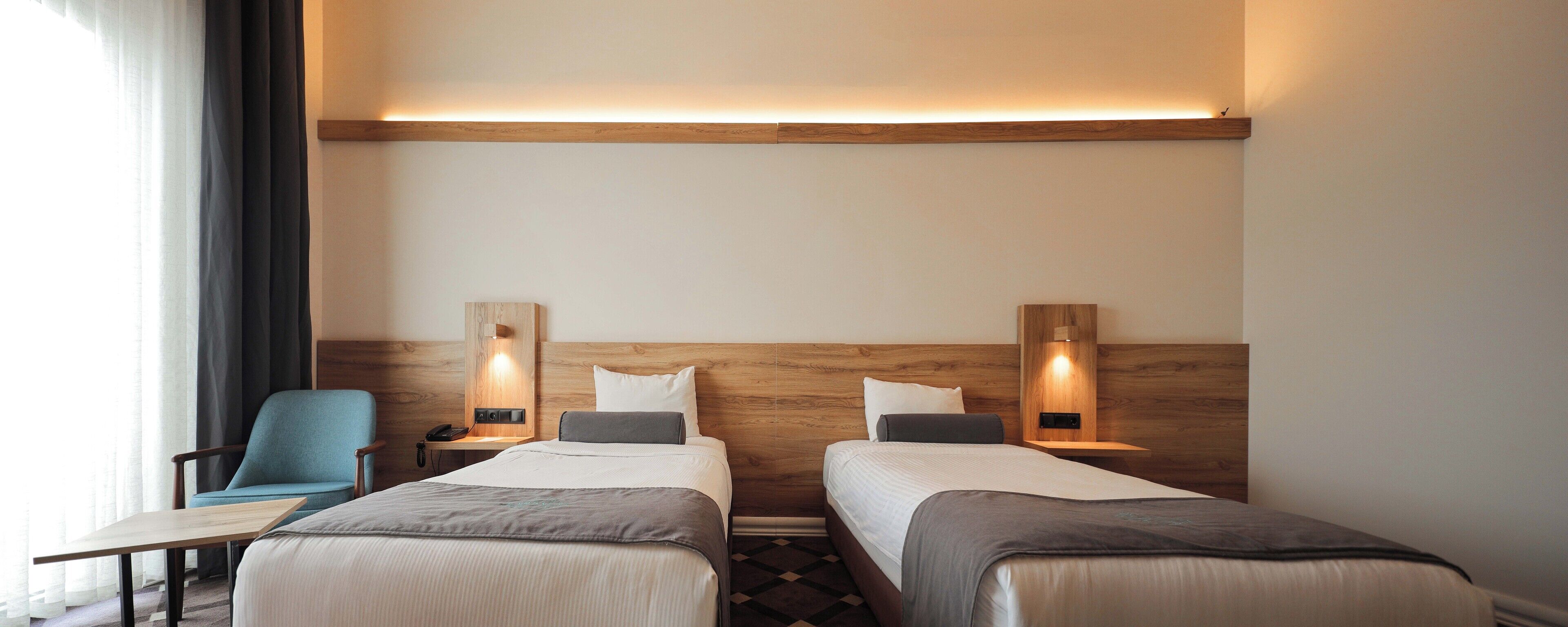 A hotel room with two beds, a desk, a chair, and a nightstand with a lamp.