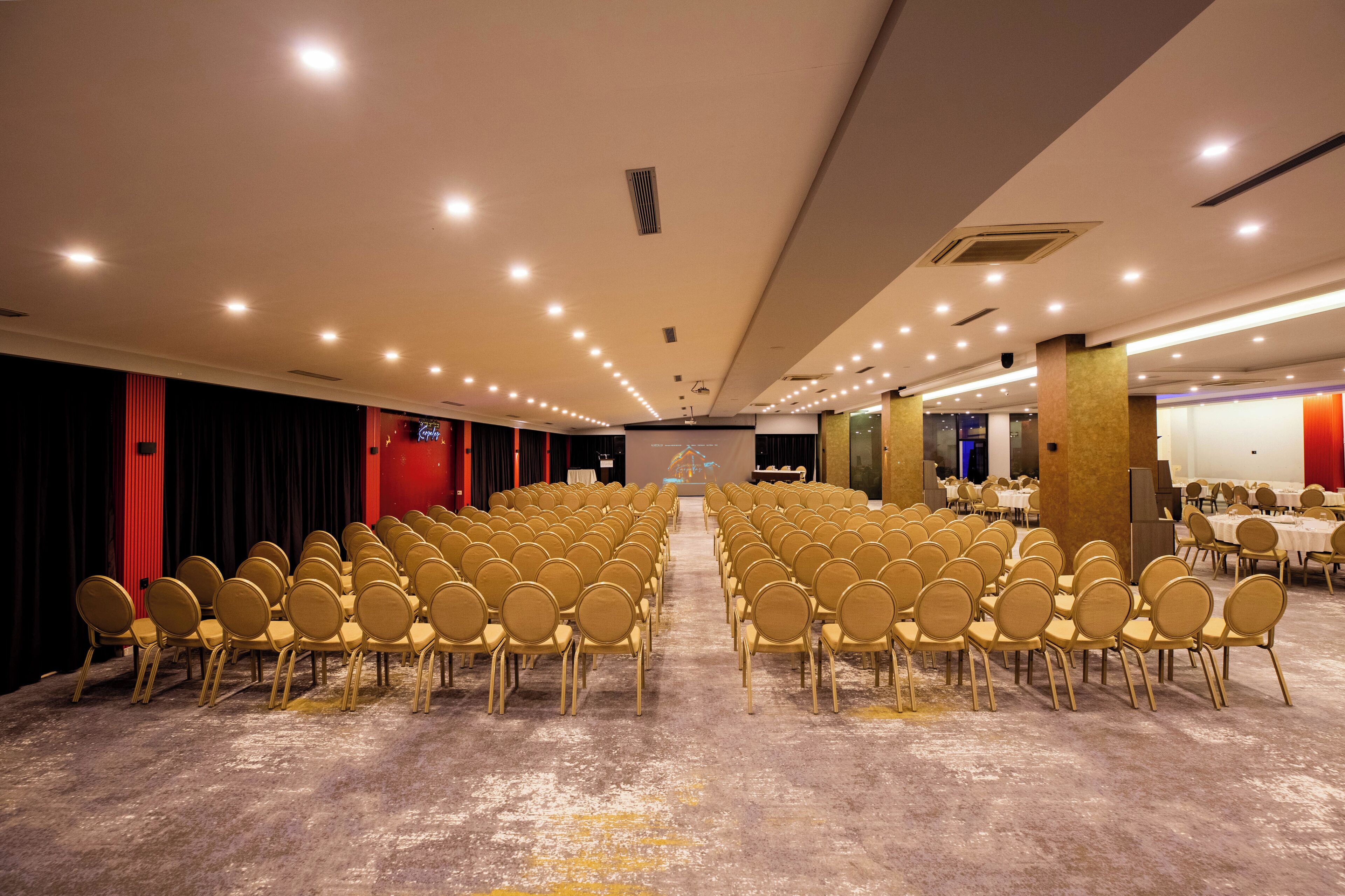 Un salón de conferencias con filas de sillas, un escenario e iluminación empotrada en el techo.
