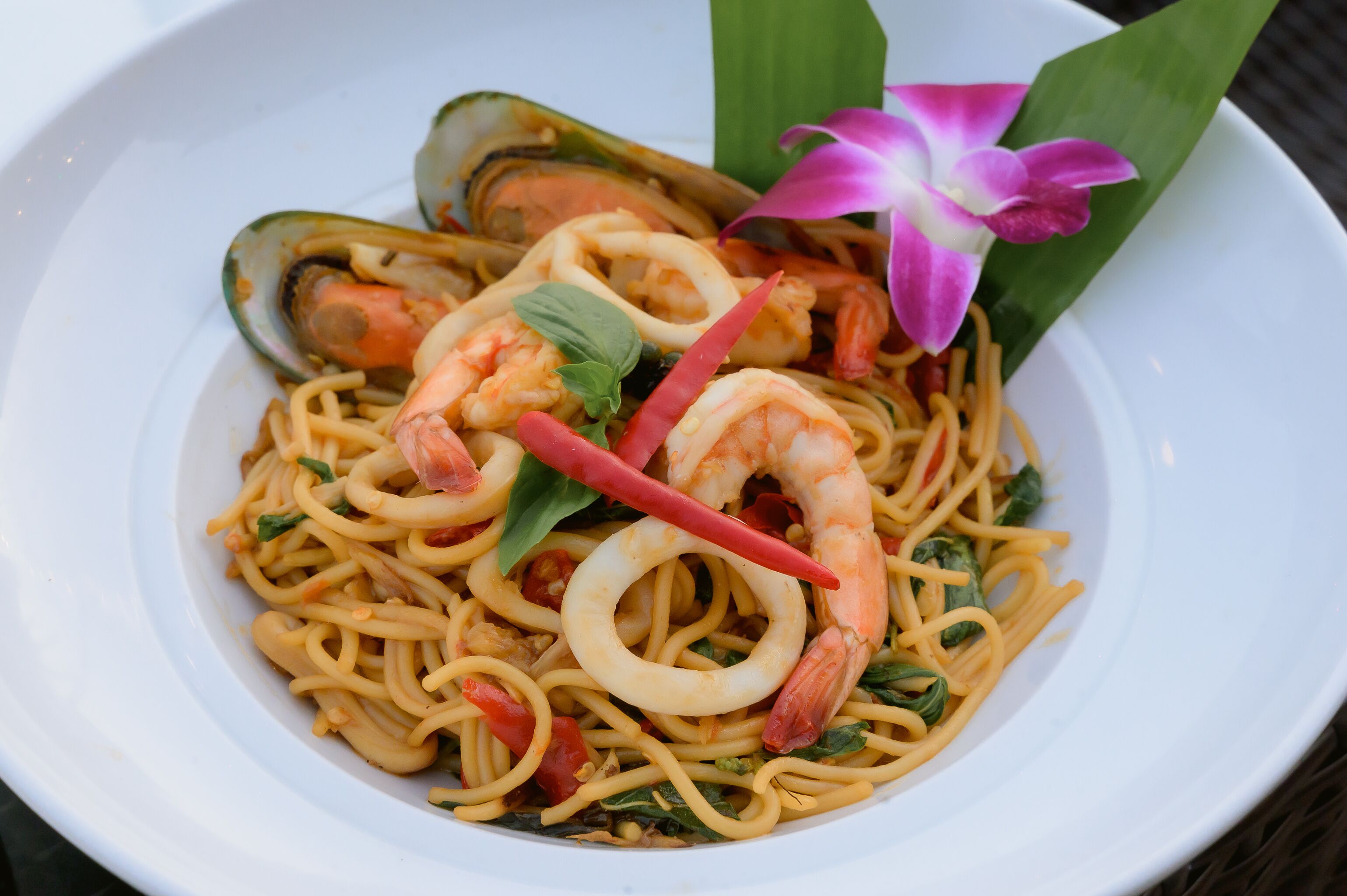A bowl of noodles with shrimp and squid, garnished with basil and a flower.