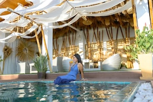 Un área de piscina con una persona relajándose, rodeada de toldos blancos, cañas de bambú y plantas en macetas.
