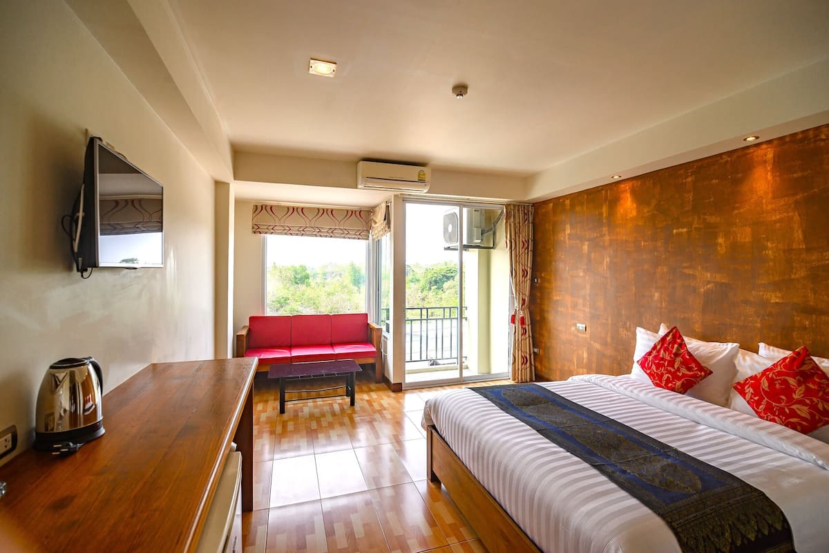 A hotel room with a bed, a wooden desk, a red sofa, and a sliding glass door leading to a balcony.